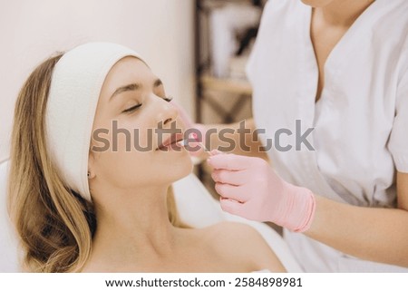 Beautician wearing pink gloves carefully applying lip mask to relaxed female client resting on treatment bed during skincare procedure