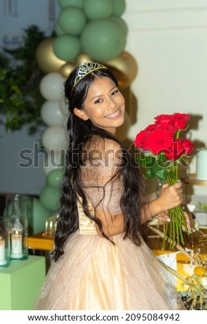 beautiful 15 year old girl celebrates her birthday with a tiara and an elegant dress and a bouquet of red roses
