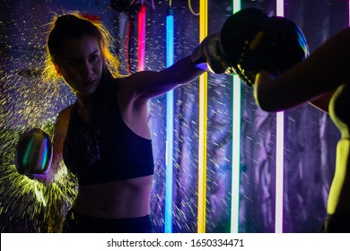 Beautiful 20s Asian Woman Trains Punching With Coach On Silver Gold Mitts Gloves. Office Girl Exercise At Modern Multi Color Fashion Neon Muay Thai Boxing Gym With Sweat Water Splash Reflect Light