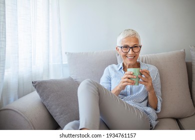 Beautiful 50 Years Old Caucasian Lady Sit Down On The Sofa Drinking Tea And Reading A Book For Afternoon Indoor Leisure Activity At Home - After Work Lifestyle For People Concept 