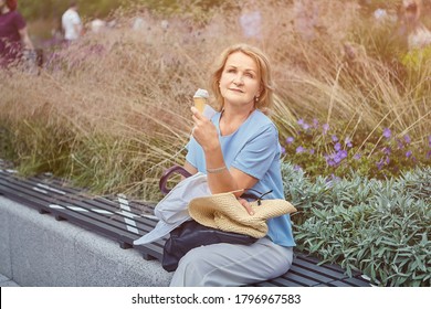 Beautiful Active Caucasian Mature Woman About 62 Years Old Is Sitting On The Bench Marked For Social Distance During Coronavirus Covid-19 Pandemic In The Public Park And Eating Ice Cream.