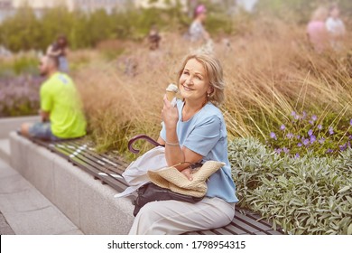 Beautiful Active Mature White Woman About 62 Years Old In Casual Cloth Is Sitting On The Bench Marked For Social Distance During Coronavirus Covid-19 Pandemic And Eating Ice Cream And Smiling.