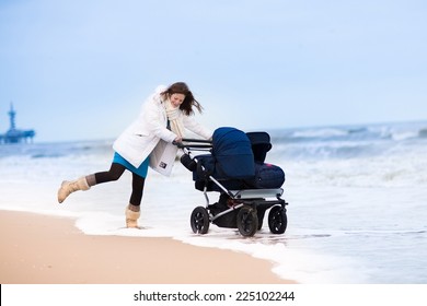 Beautiful Active Young Mother Walking On A Beach Pushing An All Terrain Double Stroller With Two Children, Baby And Toddler, On A Cold Winter Day In Holland