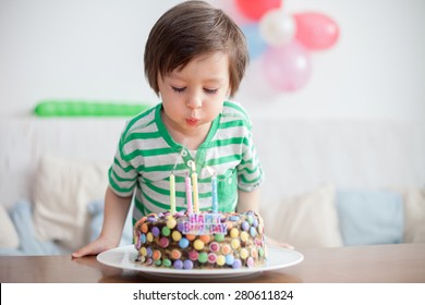 Beautiful adorable four year old boy in green shirt, celebrating his birthday, blowing candles on homemade baked cake, indoor. Birthday party for kids