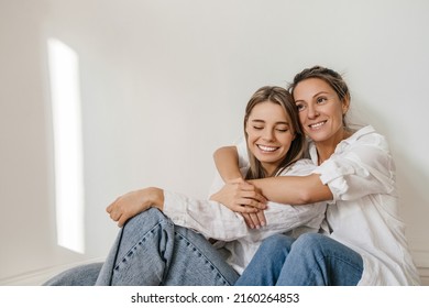 Beautiful adult caucasian woman hugs from back young girl sitting next to her on floor against wall. Blondes wear white shirts and jeans. Lifestyle, female beauty concept - Shutterstock ID 2160264853