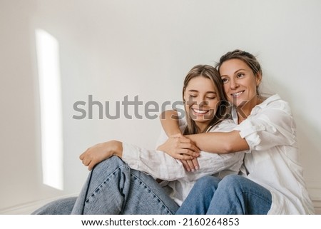 Beautiful adult caucasian woman hugs from back young girl sitting next to her on floor against wall. Blondes wear white shirts and jeans. Lifestyle, female beauty concept