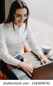 Beautiful adult caucasian woman wearing eyeglasses and dressed in white sissint at a table in coffee shop and using a laptop for working like freelancer near a big window.