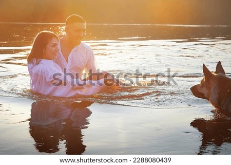 Beautiful adult couple has fun with bug dog shephers in nature in water of river or lake in summer evening at sunset. Guy and girl swim and relax with pet outdoors in clothes in white shirts and jeans