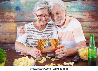 Beautiful adult mature senior white haired caucasian couple toasting with beer sitting at pub restaurant wooden table eating popcorn. Cheerful elderly people white haired having fun together