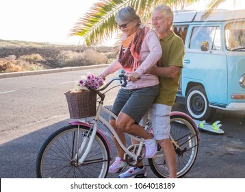 Beautiful Adult People Man And Woman Play With A Bibycle Outdoor. Old Vintage Van Camper On The Background With A Surf Table On The Ground. Alternative Lifestyle For Retired Cool