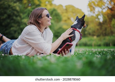 Beautiful adult woman with basenji dog resting lying on a blanket on the grass in a summer park with a smartphone - Powered by Shutterstock