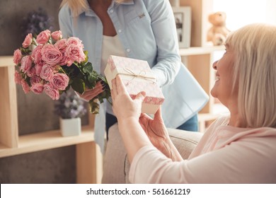 Beautiful Adult Woman Is Giving Flowers And A Gift Box To Her Mature Mother And Smiling While Sitting On Couch At Home