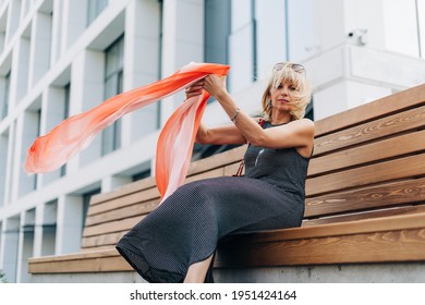 Beautiful Adult Woman With Scarf In The Wind, Sit Down On Bench Outside On A Urban City Street. Happy Lady Girl With Blonde Hair Relax Outdoors.