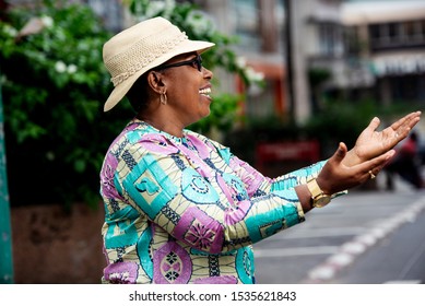 Beautiful Adult Woman Wearing A Hat Standing And Looking Sideways With Both Hands Outstretched In The Street Smiling