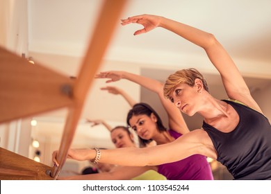 Beautiful adult women doing exercises on stretching ballet bar in Pilates class. Group of females doing yoga, pilates and fitness exercise indoors in studio.
