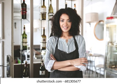 Beautiful African American barista girl in apron standing with arms folded in restaurant. Young girl with dark curly hair standing in apron at cafe. Portrait of smiling waitress wearing uniform - Powered by Shutterstock