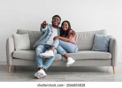 Beautiful African American black couple relaxing on sofa at home in living room, smiling man and woman spending free time together, watching TV or movie, embracing, laughing guy holding remote control