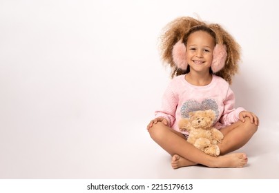 Beautiful African American Child Wearing Earmuffs Hat Hugging Teddy Bear And Smiling At Camera Isolated On White