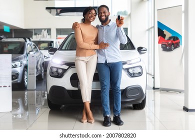 Beautiful african american couple bought car in dealership center, happy black spouses posing with keys and embracing, celebrating purchasing new family vehicle in automobile showroom, free space