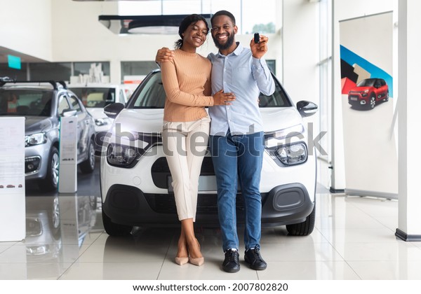 Beautiful african american couple bought car in\
dealership center, happy black spouses posing with keys and\
embracing, celebrating purchasing new family vehicle in automobile\
showroom, free space