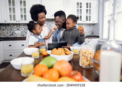 Beautiful african american family of four using digital tablet during breakfast on bright kitchen. Happy young parents watching cartoons with their two daughters while eating at home. - Powered by Shutterstock