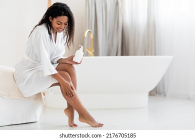Beautiful african american female applying moisturising lotion on legs while sitting in luxury bathroom interior, happy black woman in white silk robe making beauty treatments at home, copy space - Shutterstock ID 2176705185