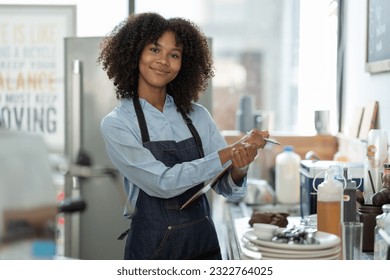 Beautiful african american female cashier or barista writing purchase details and checking availability on the clipboard at the counter in the cafe. - Powered by Shutterstock