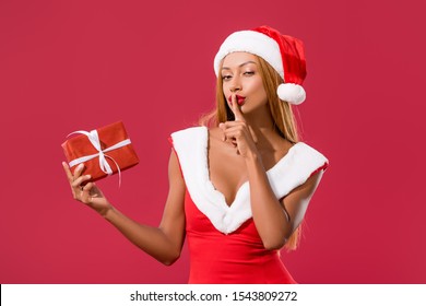 Beautiful African American Girl In Santa Hat And Christmas Dress Holding Gift Box And Showing Hush Sign Isolated On Red