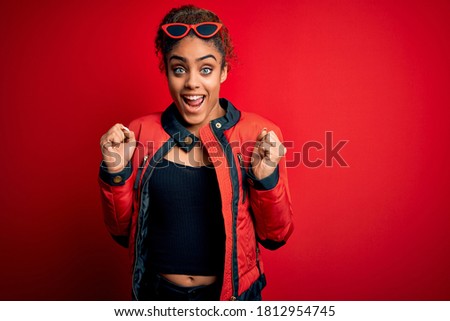 Beautiful african american girl wearing red jacket and sunglasses over isolated background celebrating surprised and amazed for success with arms raised and open eyes. Winner concept.