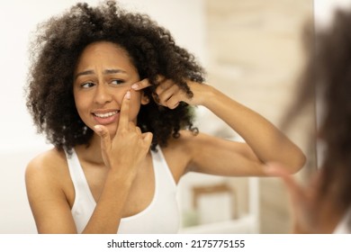 Beautiful African American Girl In White Tank Top Squeezing Pimples On Her Face While Looking At Bathroom Mirror