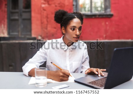 Beautiful African American girl in white shirt thoughtfully working with laptop in cozy courtyard of cafe