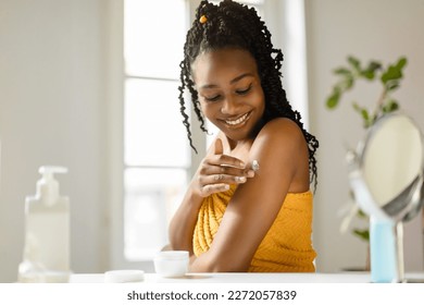 Beautiful african american lady applying cream on her shoulder, taking care of skin after bath in bedroom, free space. Young woman making daily beauty routine, pamering her body - Powered by Shutterstock