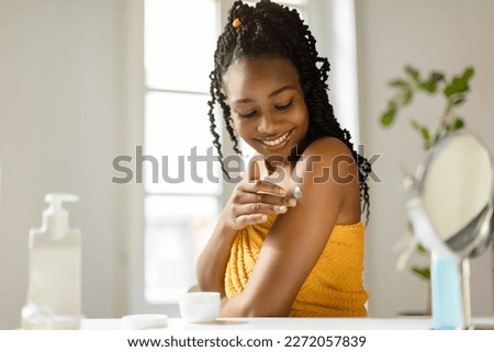 Beautiful african american lady applying cream on her shoulder, taking care of skin after bath in bedroom, free space. Young woman making daily beauty routine, pamering her body