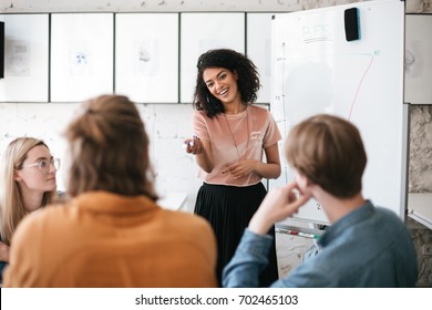 Beautiful African American Lady With Dark Curly Hair Standing Near Board And Happily Looking At Her Colleagues In Office. Young Smiling Business Woman Giving Presentation To Coworkers