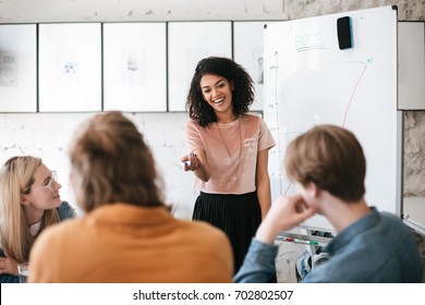 Beautiful African American lady with dark curly hair standing near board and happily discussing new project with her colleagues in office. Young smiling business woman giving presentation to coworkers - Powered by Shutterstock