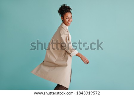 Beautiful african american lady in trench coat looking at camera. Studio shot of blithesome black young woman posing on turquoise background.