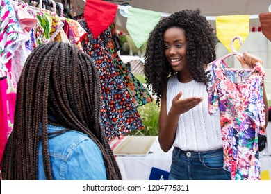 Beautiful african american market vendor presenting colorful clothes to customer at typical market - Powered by Shutterstock