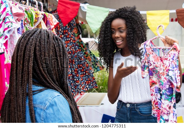 Beautiful african american\
market vendor presenting colorful clothes to customer at typical\
market