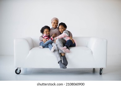 Beautiful african american muslim woman sitting on couch with her two pretty daughters and smiling on camera. Sisters embracing mother in studio. - Powered by Shutterstock