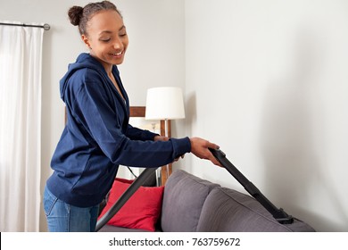 Beautiful African American Teenager Using Vacuum Cleaner On Home Sofa Fabric, Doing Cleaning Duties Chores, Smiling In Living Room Interior. Young Female Working On House Spring Cleaning, Interior.