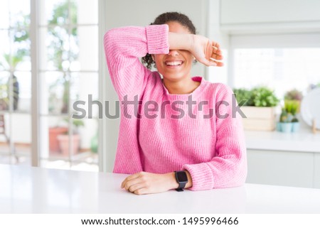 Beautiful african american woman with afro hair wearing casual pink sweater covering eyes with arm smiling cheerful and funny. Blind concept.