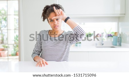Beautiful african american woman with afro hair wearing casual striped sweater making fun of people with fingers on forehead doing loser gesture mocking and insulting.