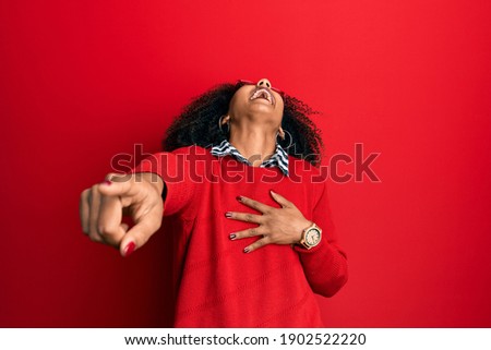 Beautiful african american woman with afro hair wearing sweater and glasses laughing at you, pointing finger to the camera with hand over body, shame expression 