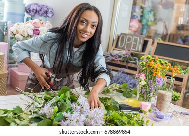 beautiful african american woman in apron holding secateurs while cutting flowers and smiling at camera in flower shop - Powered by Shutterstock