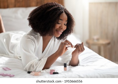 Beautiful African American Woman In Bathrobe Doing Her Nails On Bed At Home, Copy Space. Lovely Black Lady Making Manicure, Pampering Herself, Having Domestic Spa Day