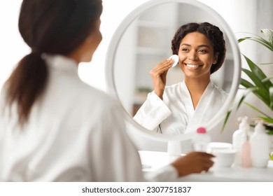 Beautiful African American Woman Cleansing Skin With Cotton Pad Near Mirror At Home, Attractive Black Female Smiling To Her Reflection, Enjoying Daily Skincare Routine, Selective Focus - Powered by Shutterstock