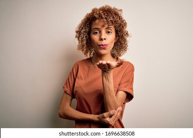 Beautiful african american woman with curly hair wearing casual t-shirt over white background looking at the camera blowing a kiss with hand on air being lovely and sexy. Love expression.