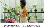 Beautiful African American woman entrepreneur working at own flower shop making bouquet and typing on laptop alone. Young female in apron makes bunch of flowers in store. Florist concept