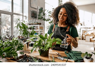 Beautiful african american woman florist small business owner in a shop sprays the leaves of a flowerpot with a sprayer, drops of water moisten the plant. Caring for indoor plants. - Powered by Shutterstock