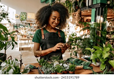 Beautiful african american woman florist in a green t-shirt and apron stands in the shop with plants on the table and scrolls the phone while working. Busy business lady taking orders online.       - Powered by Shutterstock